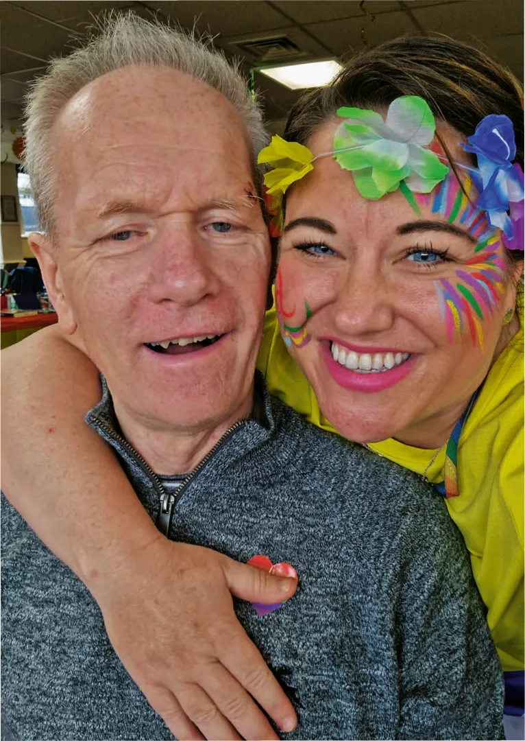 Carer with colourful face paint hugging a smiling male client during a cheerful Astar Homecare event.