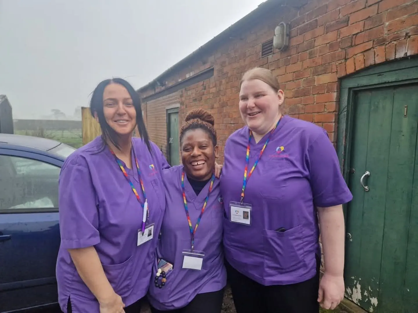 Two smiling Astar Homecare carers posing for a cheerful indoor photo in teal uniforms with rainbow lanyards.